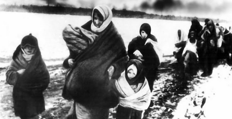 Women and children feeling Ukraine on foot through the snow in World War II with only what they could carry, as building smoke in the background.