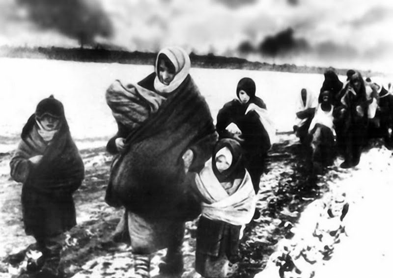 Women and children fleeing Ukraine on foot through the snow in World War II with only what they could carry, as building smoke in the background.