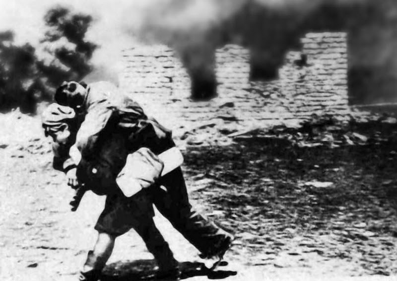 Ukrainian woman carries a man on her back, away from a smoking building in the Second World War.