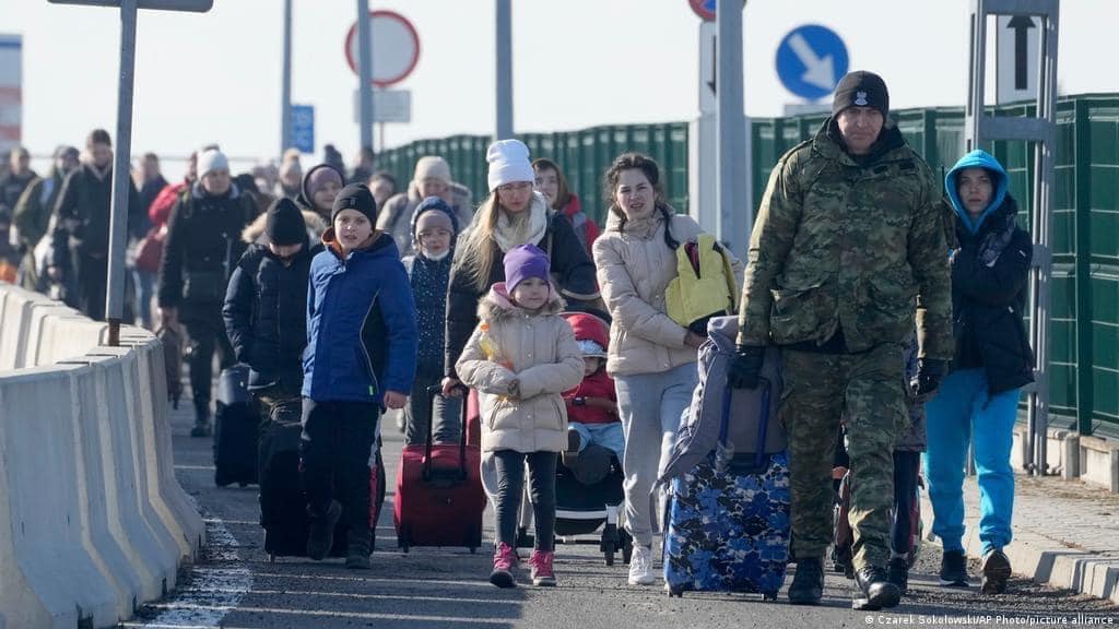 A long line of people (predominantly women and children) walk across the border from Ukraine to Poland, clad in winter gear and pulling suitcases, in March 2022..