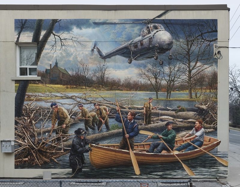 Mural showing a group of volunteers arriving in a boat at the scene of a flooded Mimico Creek in the aftermath of Hurricane Hazel. A member of the Islington Fire Brigade helps moor their boat. Behind them members of the 48th Highlanders are clearing away the debris. Above, an army supply helicopter prepares to use the 9th fairway, then high ground, as a landing strip.