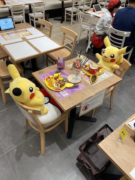 Pikachu sitting across a table from one another, with plates of treats laid out in front of them, inside the Pokemon Cafe.