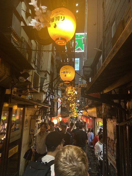 Omoide Yokocho, a narrow alley in Tokyo at night, lit by Japanese paper lanterns and filled with people, eating and shopping.