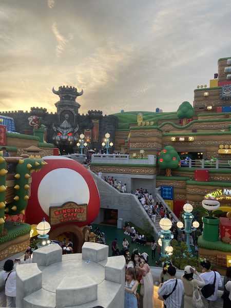 A busy scene of happy visitors exploring Super Nintendo World with Bowser's Castle and ominous skies in the background.