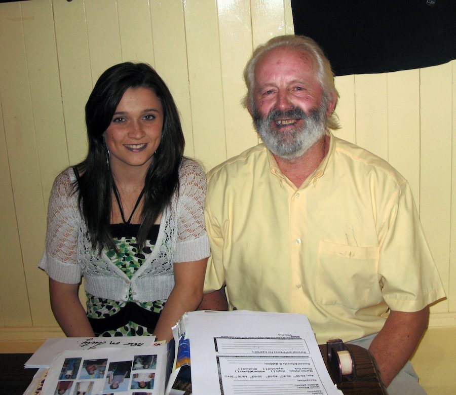 A young Irish woman dressed for a night of dancing, smiling and sitting beside a smiling Irish man in a yellow shirt with white hair, a well trimmed beard and moustache. This is Willie Daly, the Matchmaker, and his assistant, Aisling Davies.