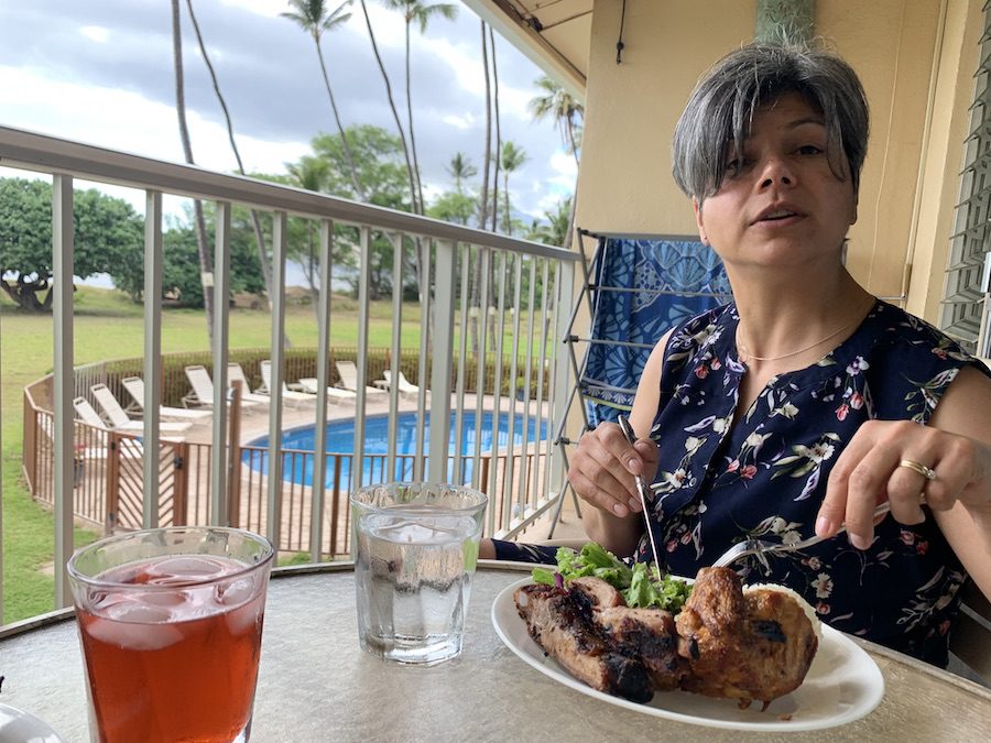 A woman on a balcony overlooking the pool and beach at Leilani Kai Resort, about to enjoy a plate of Huli Huli Chicken and Ribs.