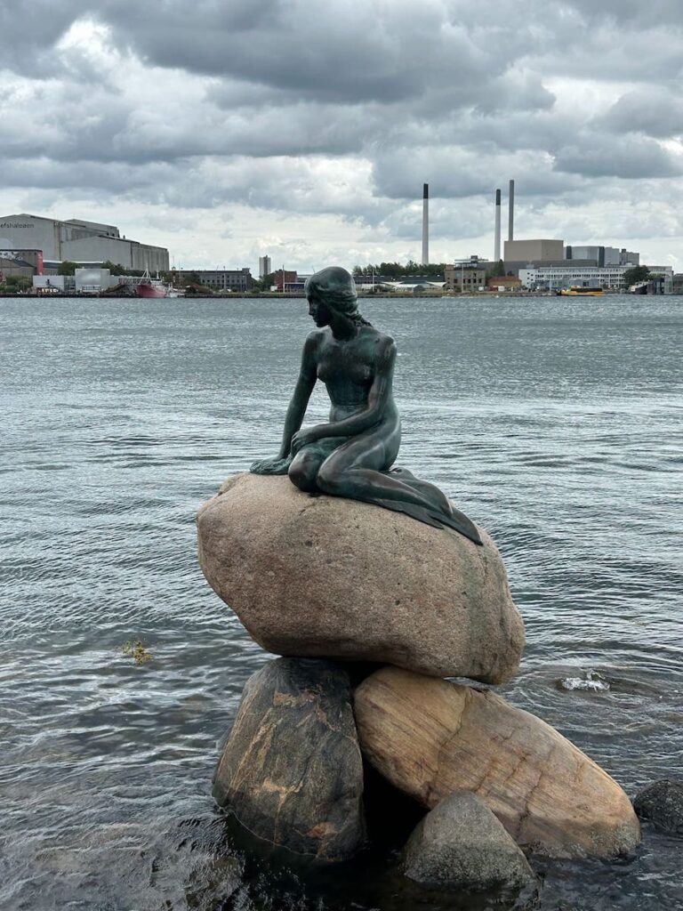 Little Mermaid sitting on the rocks over the water in Copenhagen, with smoke stacks from the factories across the water, in the background. A must stop with only 1-day in Copenhagen, Denmark.