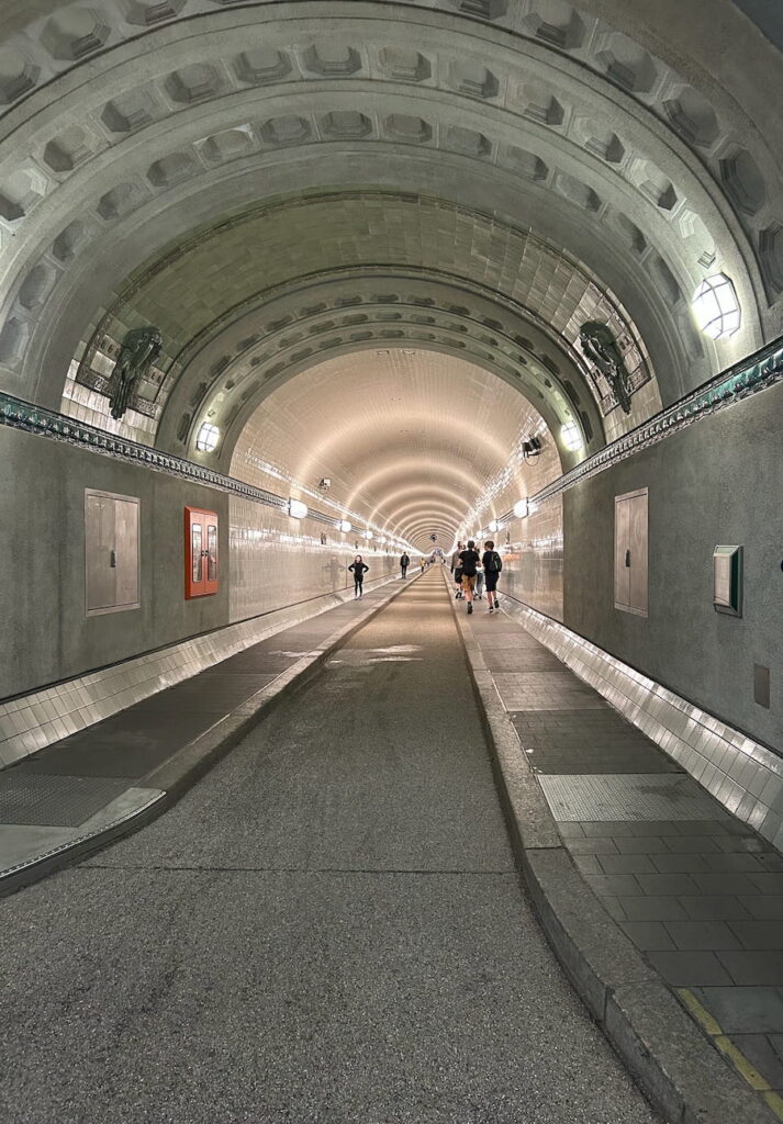 People walking through the brightly lit, white tiled Pauli Elbe Tunnel.