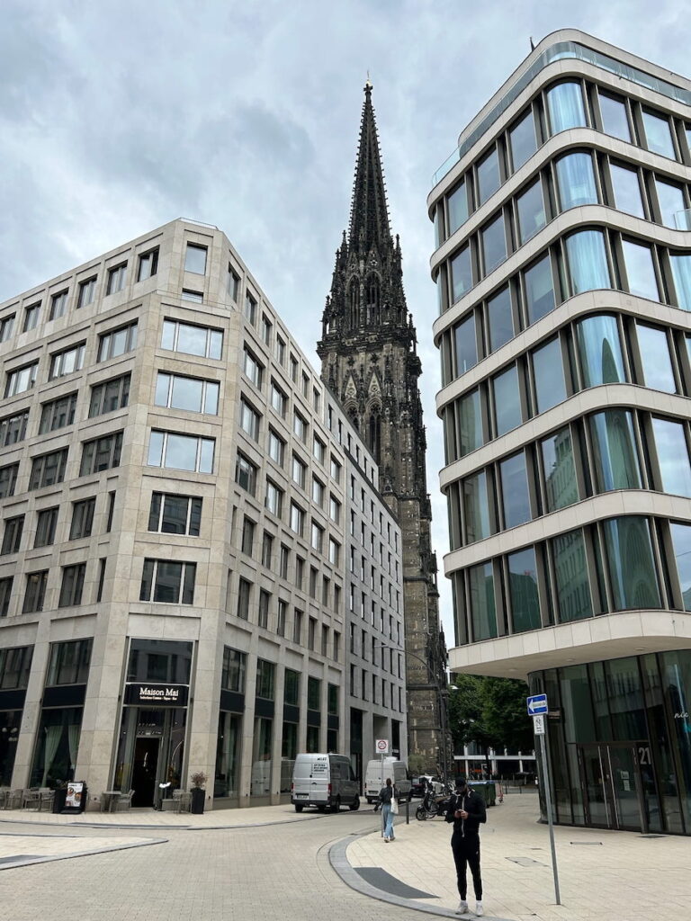 The steeple of a historic church towers over two modern buildings in Hamburg, Germany.