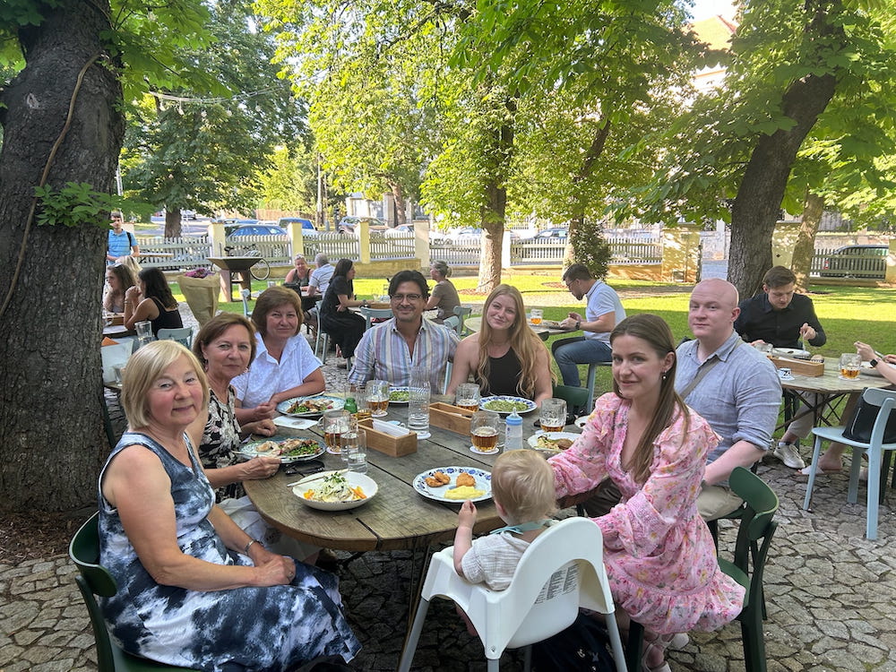 5 women, 2 men and a baby enjoying a birthday dinner around a round table outdoors on the patio at at U Mateje Restaurant in Prague, Czechia.