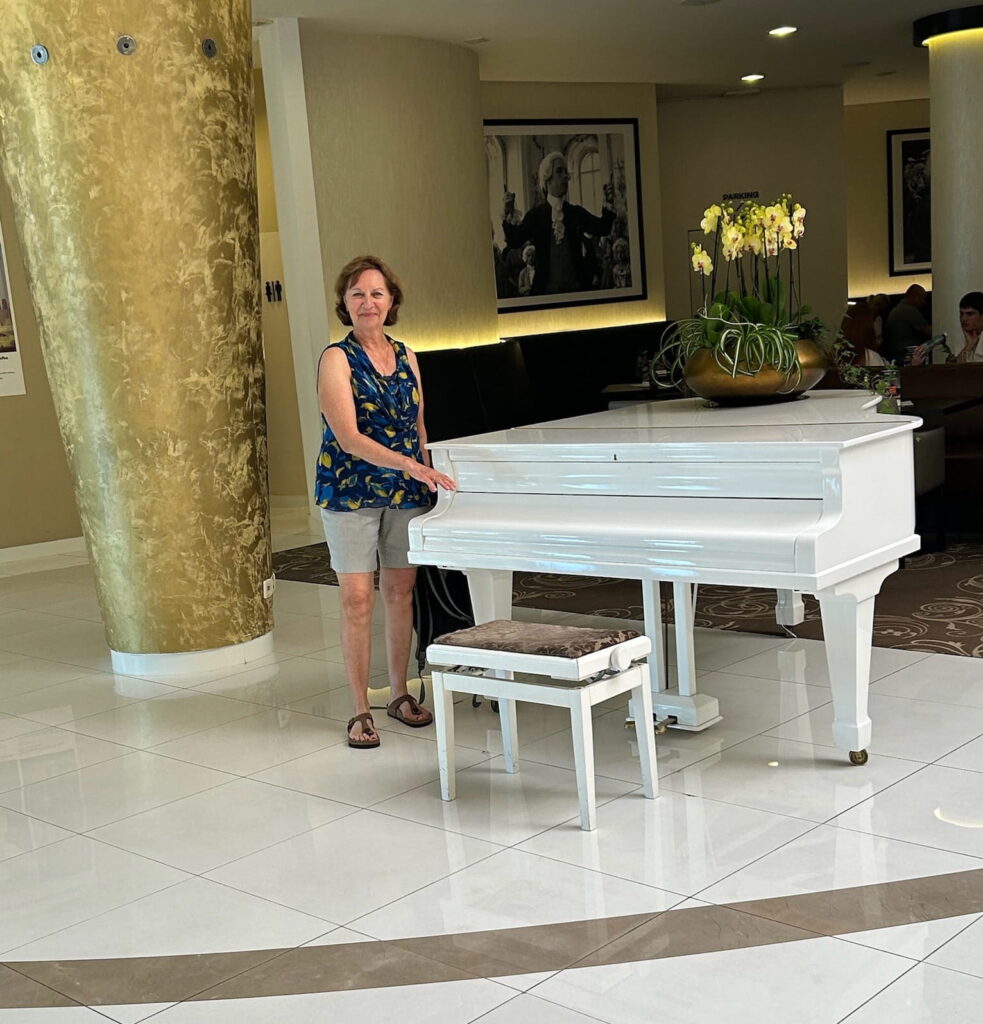 Anne Webster standing at the white piano in the lobby of the Hotel Don Giovanni in Prague, Czechia.