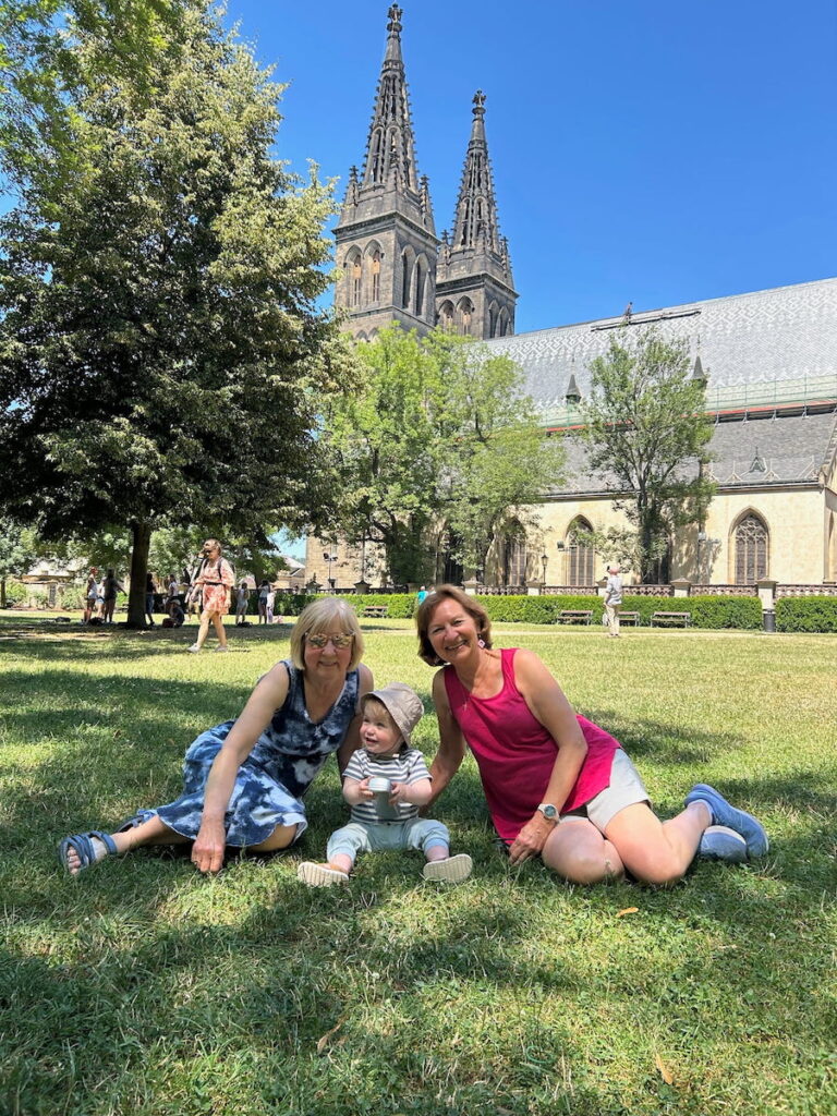 Vera, Anne and their Great Grand Nephew as a baby seat on the lawn with a church at Vysehrad in the background.